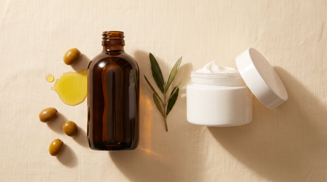 Dark amber bottle of extra virgin olive oil next to an open white moisturizer jar, with fresh olives and an olive branch, comparing olive oil skin benefits to traditional skincare products